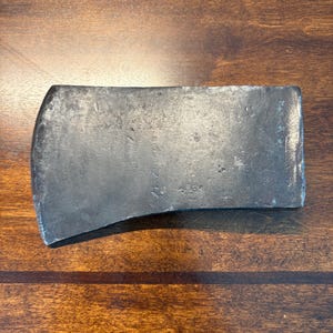 May include: A weathered, rectangular axe head with a curved cutting edge. The metal has a dark, aged appearance, showing signs of wear and use. The axe head is displayed on a wooden surface.