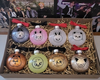 Op de afbeelding: Een doos gevuld met acht ronde, glinsterende kerstballen. Elke bal heeft een ander cartoon-diergezicht in zwart-wit. De ballen zijn gerangschikt in twee rijen van vier.