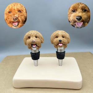 Może przedstawiać: Dwa korki do butelek w kształcie psa z brązowym futrem i uśmiechniętymi twarzami, umieszczone na kremowej podstawie. Korki mają czarną gumową uszczelkę i srebrną metalową podstawę. Dwa portrety psów są w tle.