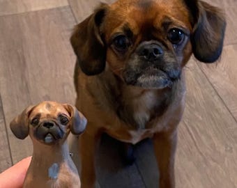 May include: A brown and black dog with short fur and big eyes sits next to a smaller brown and black dog figurine. The figurine is a replica of the larger dog.