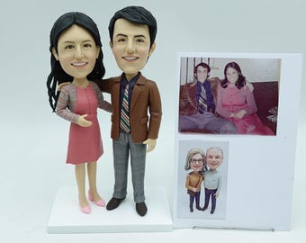 May include: A custom-made figurine set of a couple, with the woman wearing a pink dress and the man wearing a brown jacket and checked trousers. The figurines are standing on a white base. There are two photos of the couple in the background, one in colour and one in black and white.