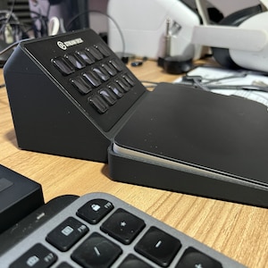 May include: A black Stream Deck with 15 buttons and a touch pad. The Stream Deck is on a wooden surface with a keyboard and a VR headset in the background.
