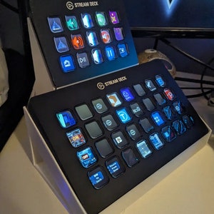 May include: Two black Stream Deck control panels with multiple illuminated buttons. The panels are on white stands. The buttons display various icons and text, including "ON", "OFF", and "MM to Inch". The Stream Deck logo is visible.