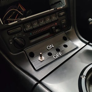 Puede incluir: Un panel de interruptor negro con un interruptor de palanca etiquetado como "ON" y "OFF". El panel está montado en el tablero de instrumentos de un automóvil.