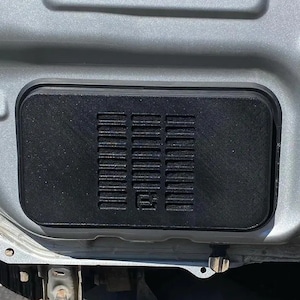 Op de afbeelding: Een close-up van een zwarte plastic ventilatiekap met een rechthoekig roosterpatroon. De ventilatiekap is gemonteerd op een zilveren carrosserie.