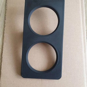 Puede incluir: Un componente rectangular negro de plástico con dos aberturas circulares. El componente parece impreso en 3D, con líneas de capa visibles. Diseñado para sujetar o enmarcar dos objetos circulares.