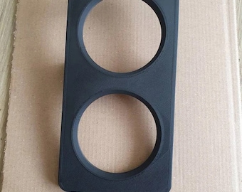 Puede incluir: Un componente rectangular negro de plástico con dos aberturas circulares. El componente parece impreso en 3D, con líneas de capa visibles. Diseñado para sujetar o enmarcar dos objetos circulares.