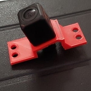 May include: A red 3D printed camera mount with a black camera attached to it.
