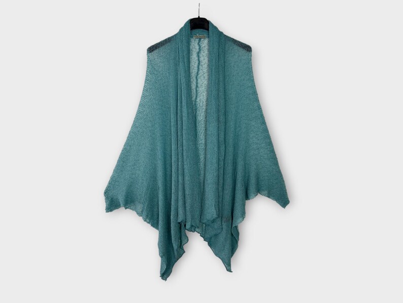 May include: A teal open-front shawl hangs on a black hanger. The lightweight fabric drapes gracefully, creating a layered effect. The shawl's design features a wide, open front and flared sleeves, ideal for layering. The colour is a muted teal.