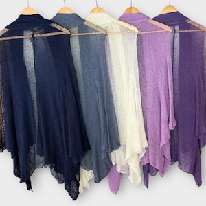 May include: Five sheer, open-front shawls in varying shades of blue, white, and purple hang on wooden hangers. The shawls have a delicate, flowing design, perfect for layering. The colors range from deep navy to light lavender.