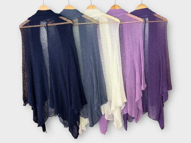 May include: Five sheer, open-front shawls in varying shades of blue, white, and purple hang on wooden hangers. The shawls have a delicate, flowing design, perfect for layering. The colors range from deep navy to light lavender.