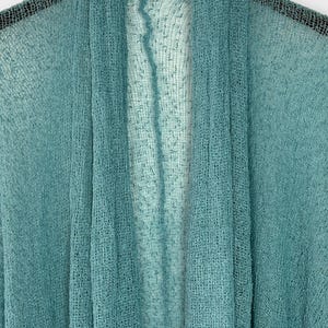 May include: A teal open-front shawl made from a sheer, woven material. The shawl is draped, highlighting its lightweight and airy texture. The colour transitions from a darker teal to a lighter shade, creating a subtle ombre effect. A versatile accessory.