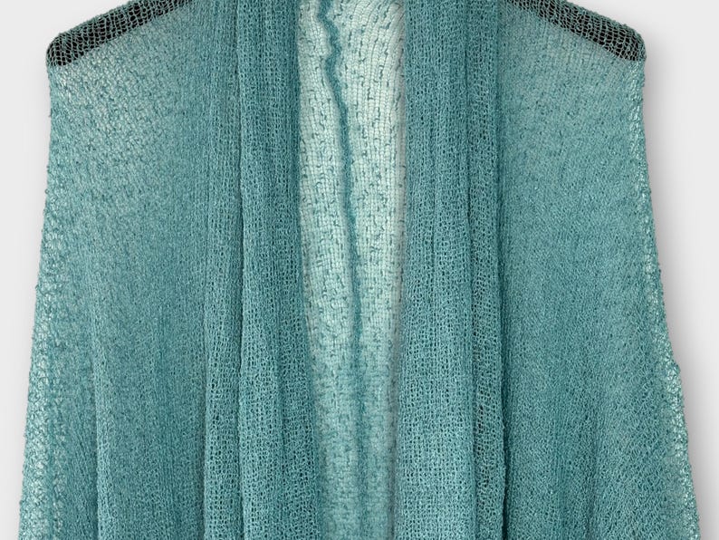 May include: A teal open-front shawl made from a sheer, woven material. The shawl is draped, highlighting its lightweight and airy texture. The colour transitions from a darker teal to a lighter shade, creating a subtle ombre effect. A versatile accessory.