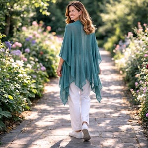 May include: A woman wearing a teal poncho and white trousers walks along a stone path in a garden. The poncho has a layered design and drapes loosely. The path is lined with flowers and greenery, creating a natural, outdoor setting.