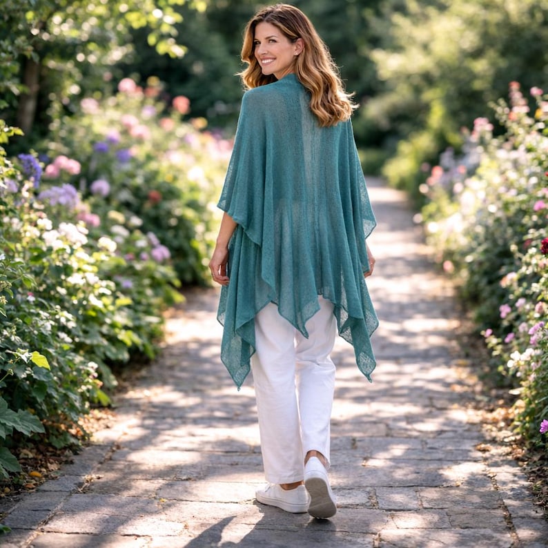 May include: A woman wearing a teal poncho and white trousers walks along a stone path in a garden. The poncho has a layered design and drapes loosely. The path is lined with flowers and greenery, creating a natural, outdoor setting.