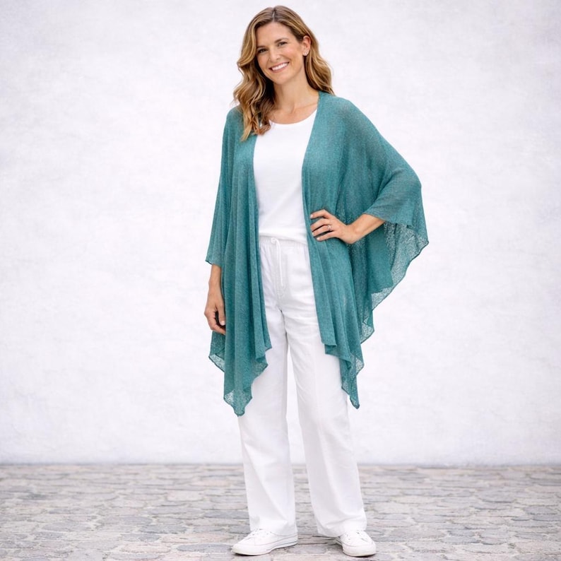 May include: A teal open-front cardigan with a draped design. The cardigan is worn with a white top and white trousers. The outfit is completed with white trainers. The overall style is casual and comfortable.