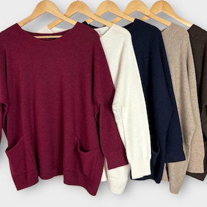 Op de afbeelding: Een rij van vijf truien met lange mouwen in verschillende kleuren: bordeauxrood, gebroken wit, marineblauw, beige en bruin. Elke trui heeft een ronde hals en twee voorzakken. De truien hangen aan houten kleerhangers.