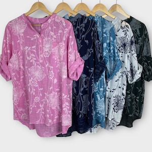 Könnte beinhalten: Fünf Hemden mit floralem Muster in verschiedenen Farben, darunter Rosa, Marineblau, Blau, Weiß und Dunkelgrün. Jedes Hemd hat einen Stehkragen und hochgekrempelte Ärmel. Die Hemden hängen an Holzbügeln vor einem schlichten Hintergrund.