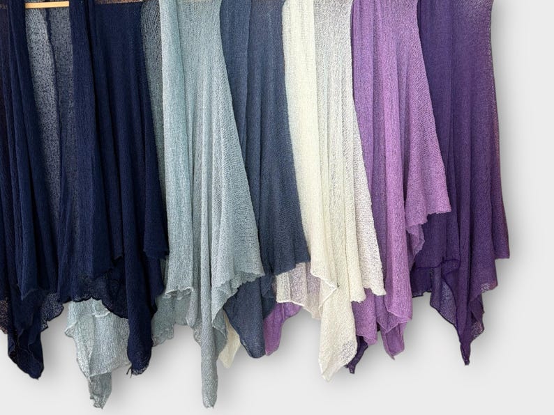 May include: A collection of lightweight, open-front cardigans in various colors, including navy, light blue, white, lavender, and deep purple. The cardigans are displayed hanging, showcasing their flowing, draped design. The fabric appears to be a sheer knit.