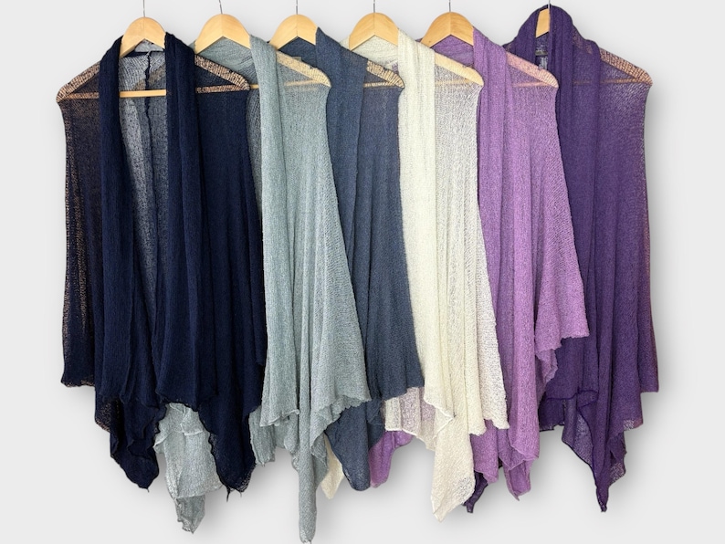 May include: A collection of open-front cardigans in various colors, including navy, gray, off-white, and purple, hanging on wooden hangers. The cardigans have a lightweight, sheer texture and a draped design.