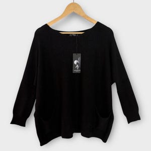 May include: A black sweater with long sleeves and two front pockets hangs on a wooden hanger. The sweater has a scoop neckline and a tag with a logo. The garment is made of a soft material.
