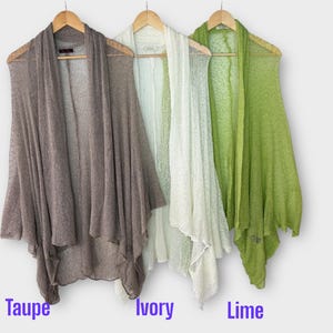 May include: Three open-front cardigans in taupe, ivory, and lime green hang on wooden hangers. The lightweight, sheer fabric drapes loosely. The colors are labeled below the cardigans.