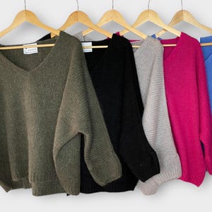 May include: A collection of five long-sleeved V-neck sweaters hanging on wooden hangers, displayed in olive green, black, light gray, magenta, and blue. Each sweater features a relaxed fit with ribbed cuffs and a visible white brand label.