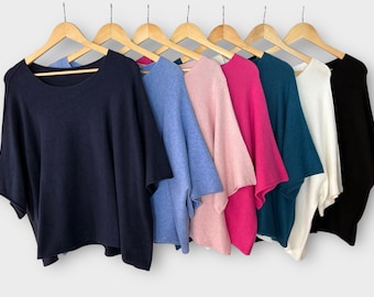 May include: A collection of seven short-sleeved knit sweaters hanging on wooden hangers. The sweaters are displayed in a row, featuring a variety of solid colors including navy blue, light blue, soft pink, vibrant magenta, teal, white, and black.