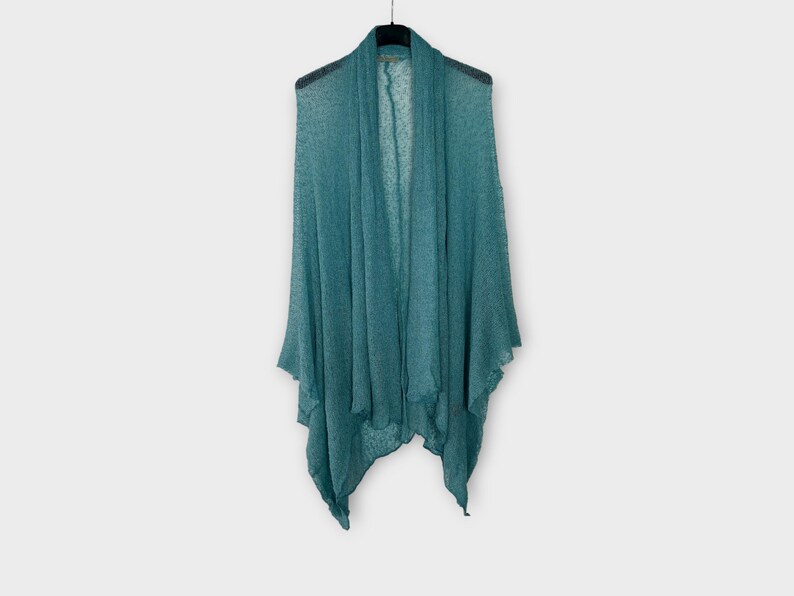 May include: A teal shawl hangs on a black hanger against a white backdrop. The shawl features a draped design with a flowing, open front. The fabric looks lightweight and slightly textured, with a subtle sheen. The garment is a wrap.