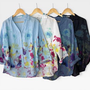 May include: Four long-sleeved button-down shirts hanging on wooden hangers. The shirts feature a watercolour-style floral print in shades of blue, yellow, pink, and purple. The shirts are available in light blue, white, medium blue, and black fabric.