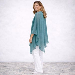 May include: A teal-coloured, lightweight poncho with a flowing design. The poncho is worn with white trousers and trainers. The fabric appears to be semi-sheer, with a subtle texture. The overall style is casual and comfortable.