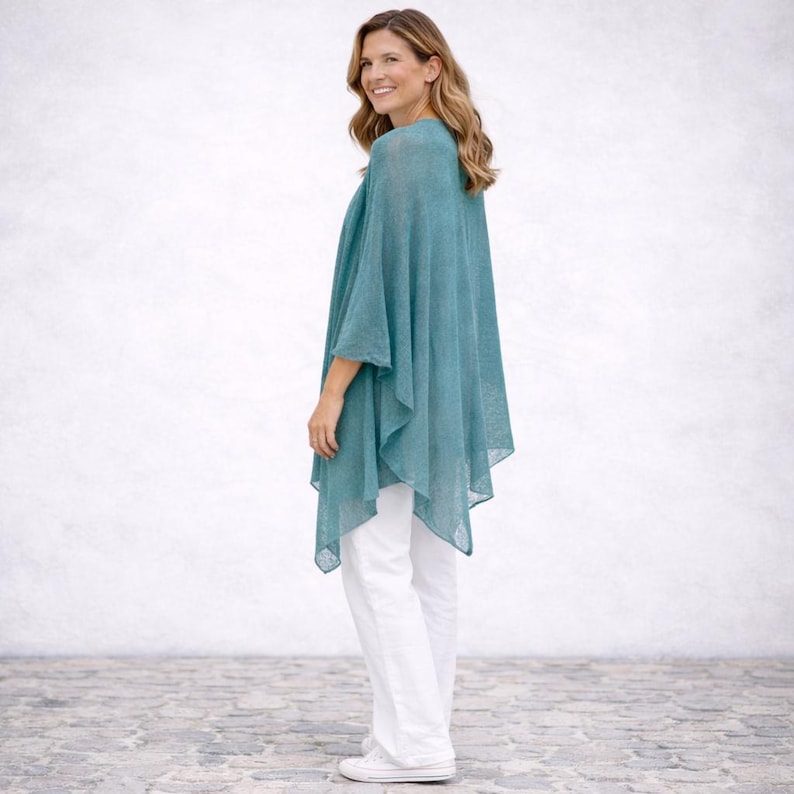 May include: A teal-coloured, lightweight poncho with a flowing design. The poncho is worn with white trousers and trainers. The fabric appears to be semi-sheer, with a subtle texture. The overall style is casual and comfortable.