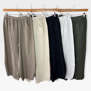 May include: Five pairs of linen trousers in shades of taupe, beige, black, white, and olive green. Each pair features an elasticated waistband and side pockets, displayed on a wooden rail.