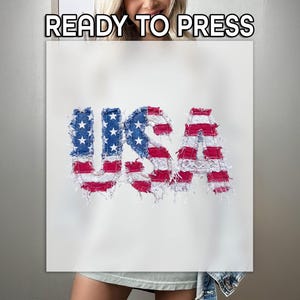 May include: A white t-shirt featuring the text USA in a distressed, frayed fabric style. The letters are patterned with the American flag, displaying blue sections with white stars and red and white horizontal stripes. The text READY TO PRESS is visible.