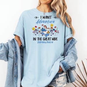 May include: Light blue t-shirt with the text "I WANT ADVENTURE IN THE GREAT WIDE somewhere". The shirt features a graphic of a woman reading surrounded by colorful flowers. The shirt is a casual style.