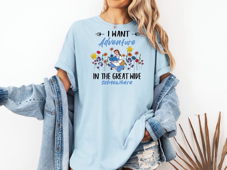 May include: Light blue t-shirt with the text "I WANT ADVENTURE IN THE GREAT WIDE somewhere". The shirt features a graphic of a woman reading surrounded by colorful flowers. The shirt is a casual style.