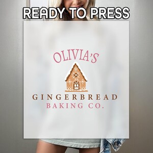 May include: A white t-shirt with a pink and brown logo that says "Olivia's Gingerbread Baking Co." and features a gingerbread house illustration.