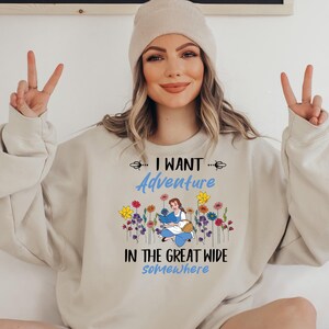 May include: Beige sweatshirt with the text "I WANT Adventure IN THE GREAT WIDE somewhere". The design features a cartoon illustration of a woman reading a book surrounded by colorful flowers.