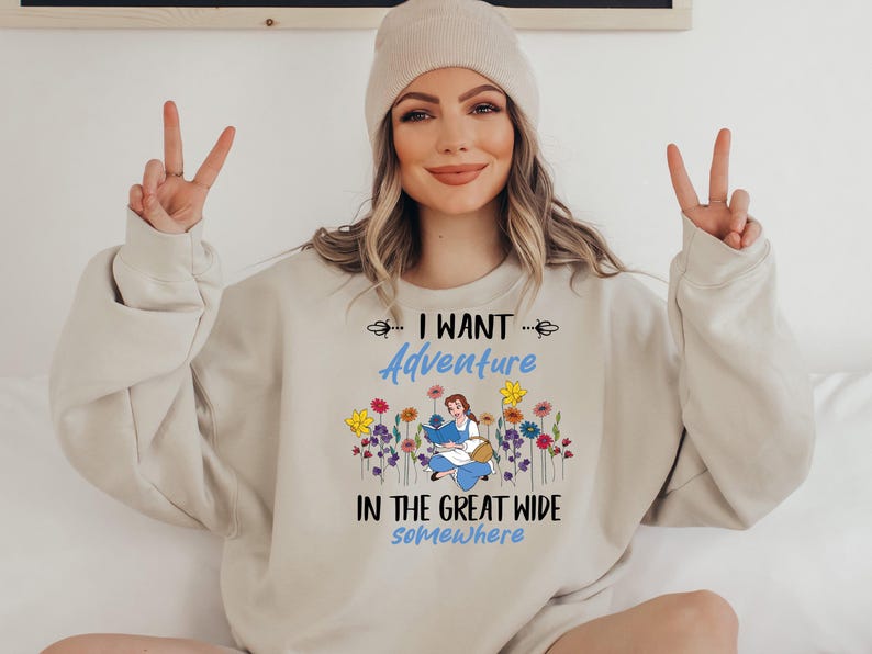 May include: Beige sweatshirt with the text "I WANT Adventure IN THE GREAT WIDE somewhere". The design features a cartoon illustration of a woman reading a book surrounded by colorful flowers.