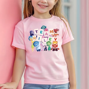 May include: A light pink t-shirt featuring the phrase "TEAM RILEY" with cartoon characters from the movie Inside Out. The shirt is short-sleeved and worn by a child. The background is a light pink wall.
