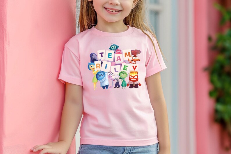 May include: A light pink t-shirt featuring the phrase "TEAM RILEY" with cartoon characters from the movie Inside Out. The shirt is short-sleeved and worn by a child. The background is a light pink wall.