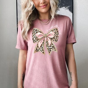 May include: A pink t-shirt with a large pink and leopard print bow with gold accents. The text "it is well with my soul" is printed on the shirt.