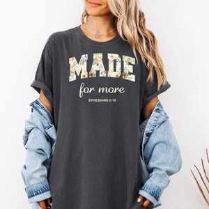 Op de afbeelding: Een donkergrijs T-shirt met een bloemenprint met de tekst "MADE for more" en "EPHESIANS 2:10" in wit.