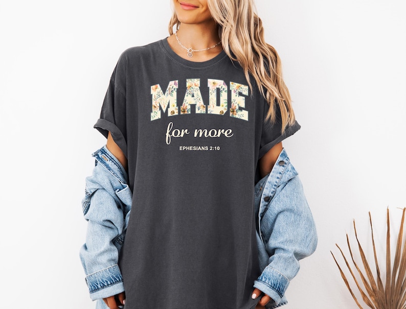 Op de afbeelding: Een donkergrijs T-shirt met een bloemenprint met de tekst "MADE for more" en "EPHESIANS 2:10" in wit.