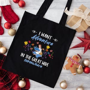 May include: Black tote bag with the text "I WANT Adventure IN THE GREAT WIDE somewhere". The bag features a colorful illustration of a woman reading a book surrounded by flowers. Christmas decorations are in the background.