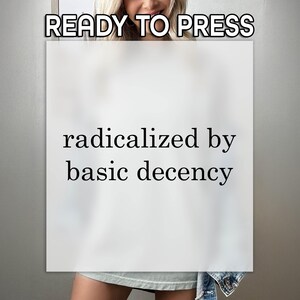 May include: A white rectangular sign with the text "READY TO PRESS" at the top and "radicalized by basic decency" in a black font. The sign is held in front of a person.
