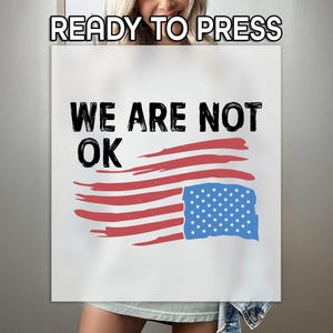 May include: White rectangular sign with the text "READY TO PRESS" at the top. Below, the words "WE ARE NOT OK" are in black, above a stylized American flag with red stripes and a blue star field.