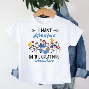 May include: White t-shirt with the text "I WANT Adventure IN THE GREAT WIDE somewhere". The shirt features a cartoon illustration of a girl reading a book surrounded by colorful flowers. The shirt is hanging on a wooden hanger.