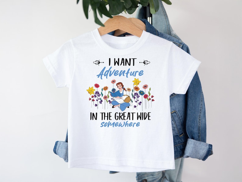 May include: White t-shirt with the text "I WANT Adventure IN THE GREAT WIDE somewhere". The shirt features a cartoon illustration of a girl reading a book surrounded by colorful flowers. The shirt is hanging on a wooden hanger.