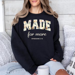 Op de afbeelding: Zwart sweatshirt met bloemenlettertype met de tekst "MADE for more" en de Bijbeltekst Efezi&euml;rs 2:10.